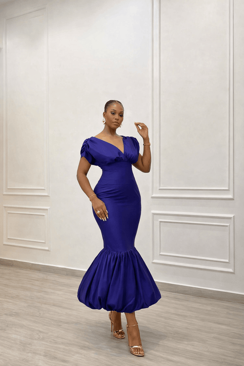 Woman wearing a blue dress standing against a white paneled wall.